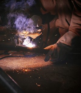 A machine operator working on a galvanized steel welding project while smoke and fumes rise from the source.