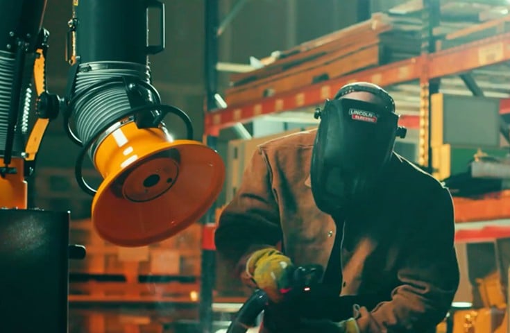 Welder operating a portable fume extractor with an articulating extraction arm positioned at the weld point inside an industrial fabrication shop.